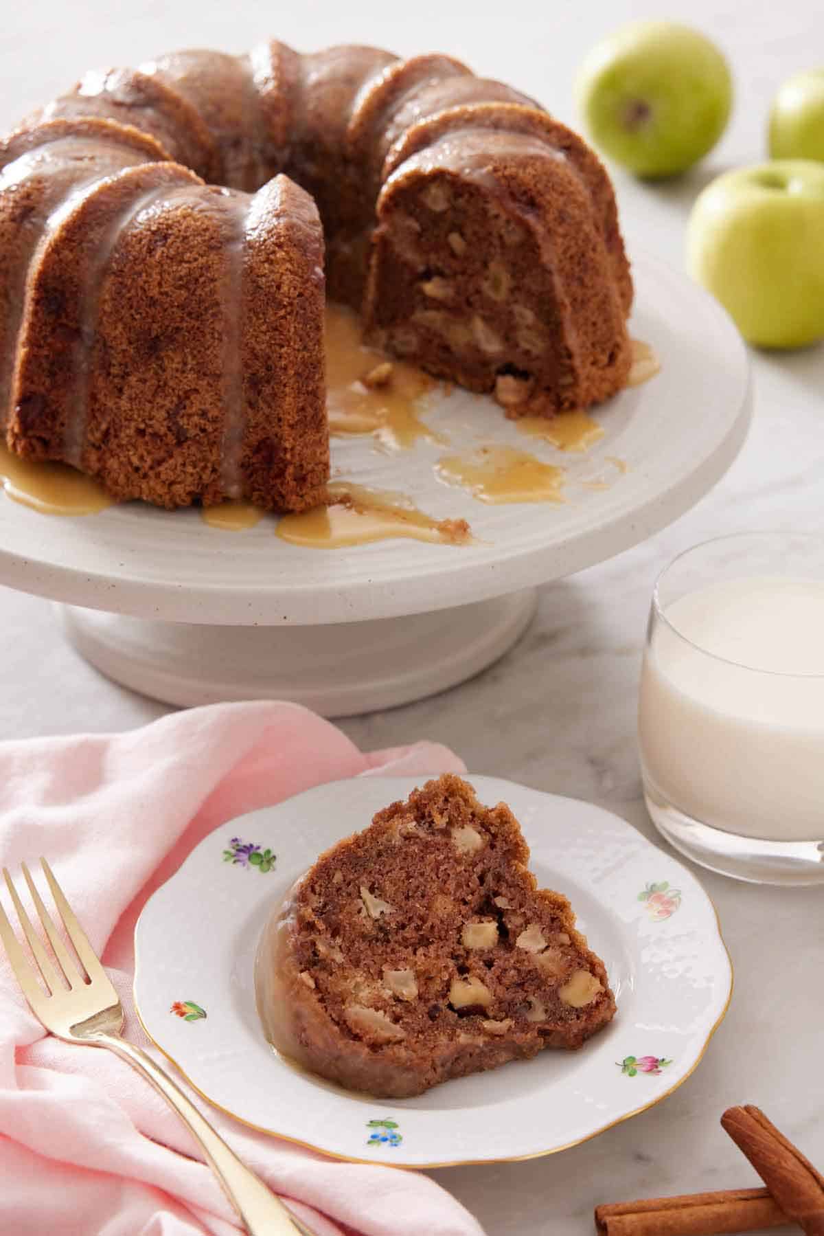 A plate with a slice of apple bundt cake with a cake stand in the background holding the rest of the cake.