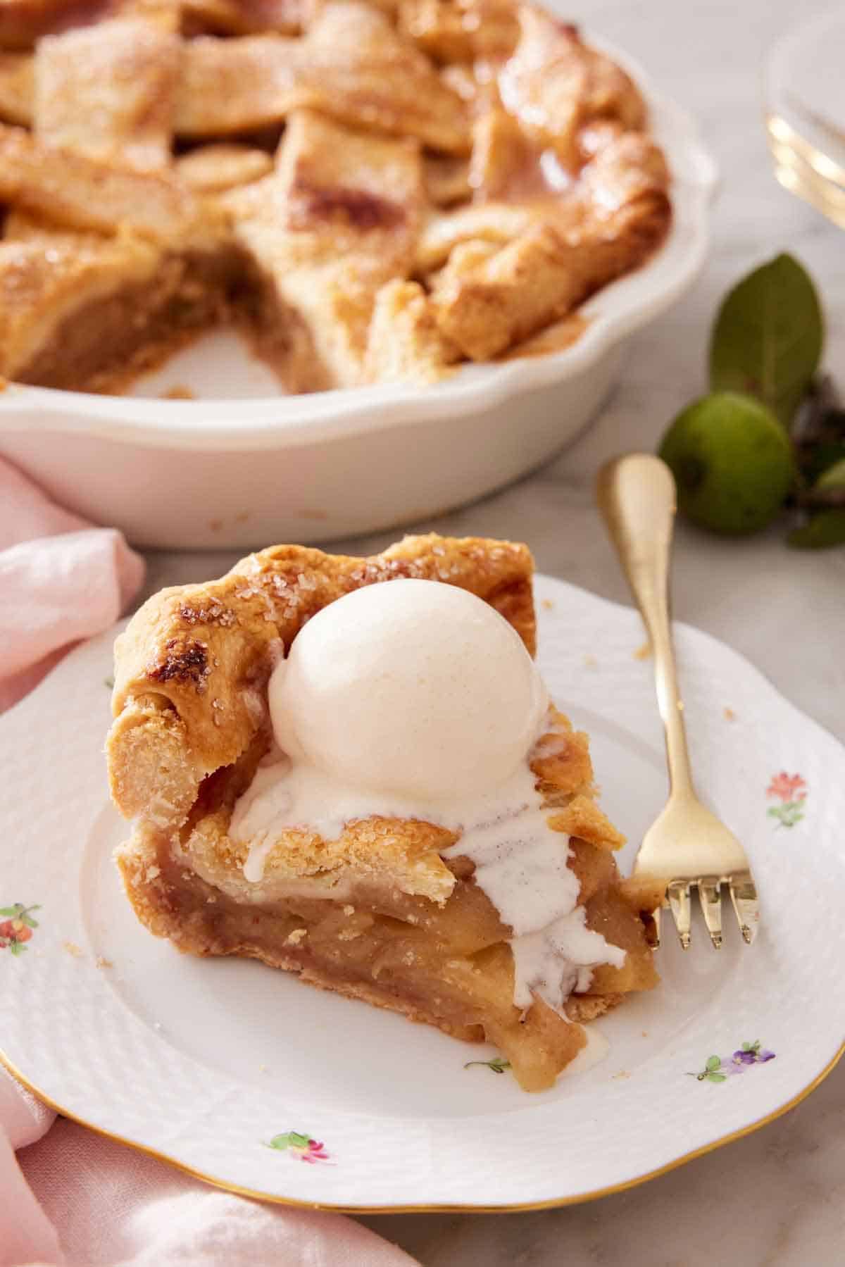 A plate with a slice of apple pie with a scoop of melting vanilla ice cream on top. A fork beside the slice and the rest of the pie in the background.