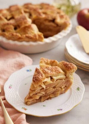 A slice of apple pie on a plate in front of the rest of the pie.