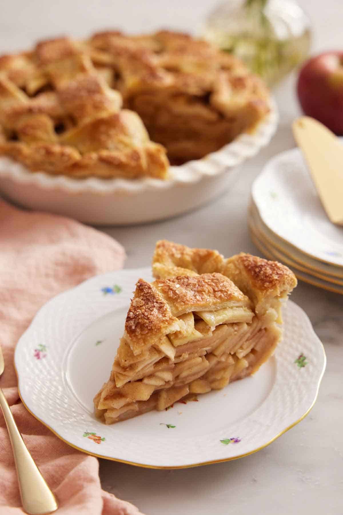 A slice of apple pie on a plate in front of the rest of the pie.