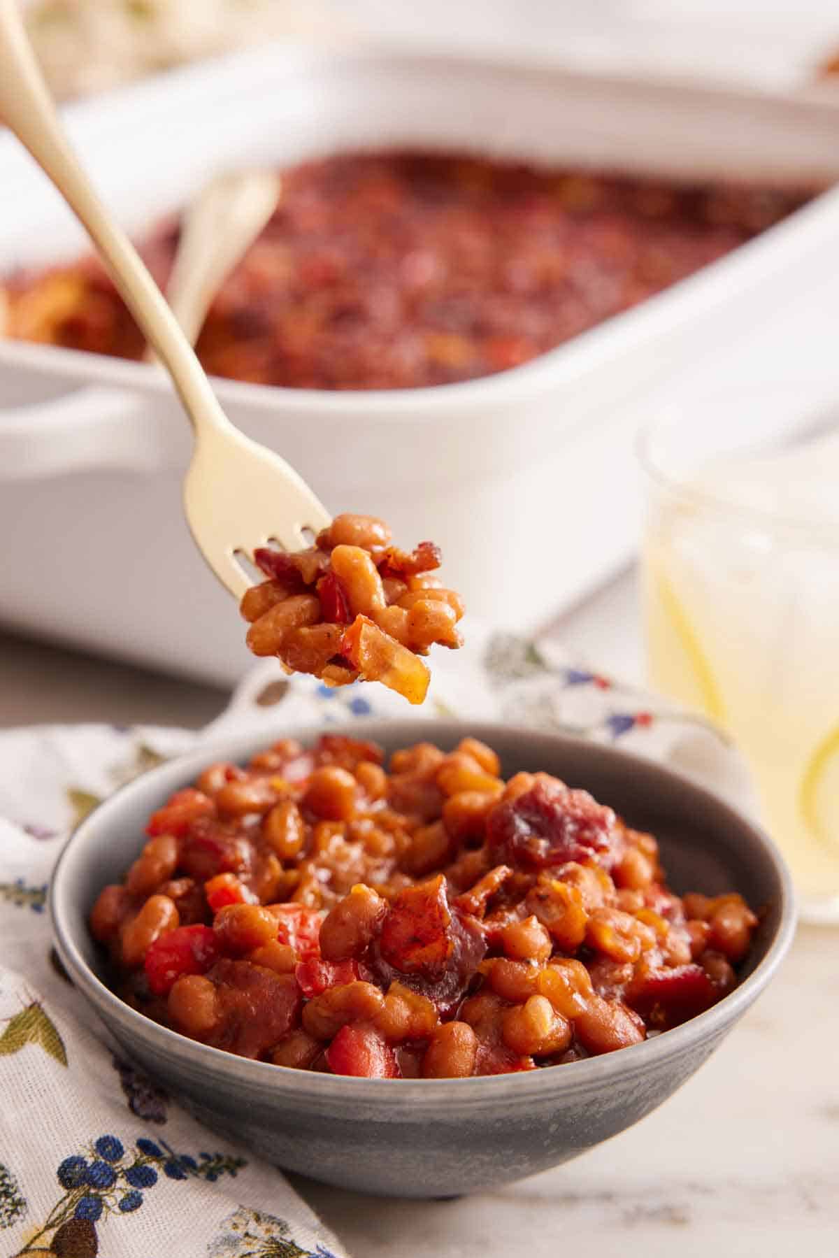 A forkful of baked beans lifted from a bowl with a baking dish in the background.