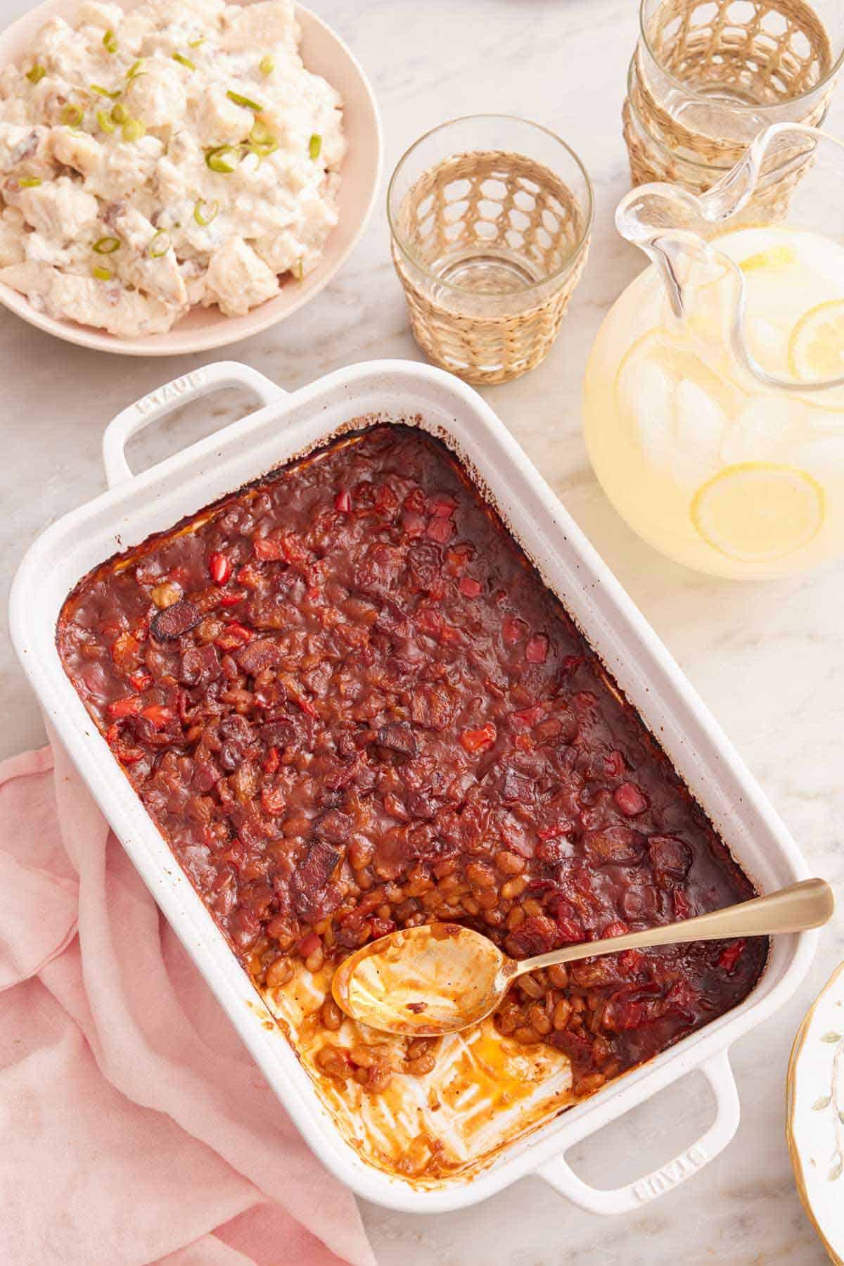 Overhead view of a baking dish with baked beans with a serving scooped out with the spoon still inside. Lemonade, glasses, and potato salad off to the side.