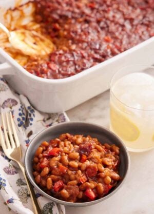 A bowl of baked beans with a glass of lemonade and baking dish of baked beans in the background.