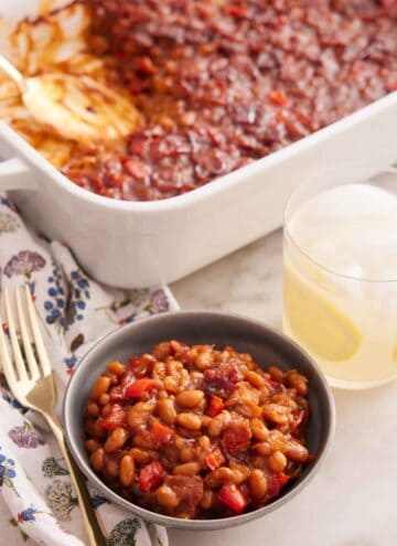 A bowl of baked beans with a glass of lemonade and baking dish of baked beans in the background.