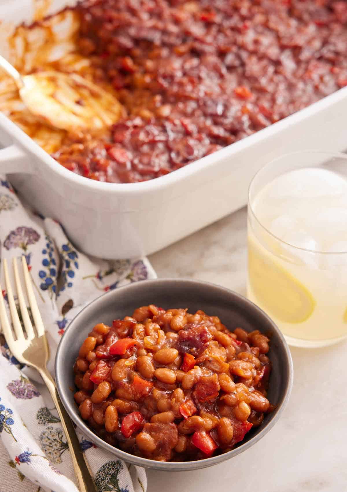 A bowl of baked beans with a glass of lemonade and baking dish of baked beans in the background.
