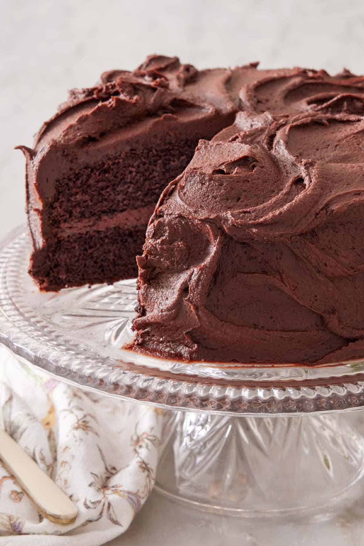 A chocolate cake with a slice taken out on a clear cake stand.