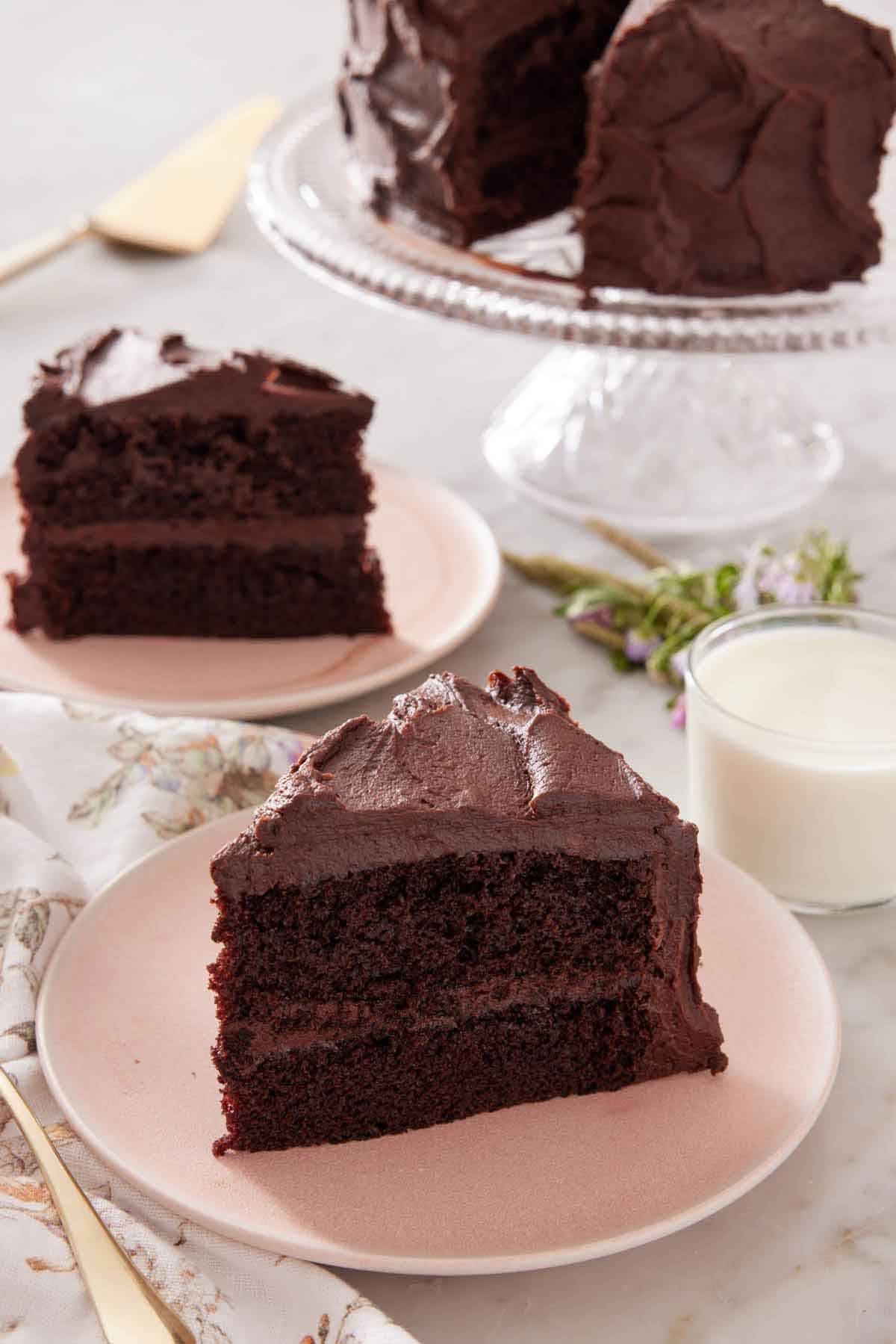 Two plates with sliced chocolate cake along with a glass of milk and cake stand with more cake.
