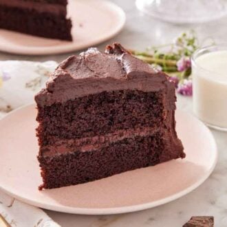 A slice of chocolate cake with a glass of milk in the back along with some flowers, a fork, and linen napkin.