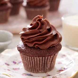 A chocolate cupcake on a plate with frosting on top and a glass of milk in the back.