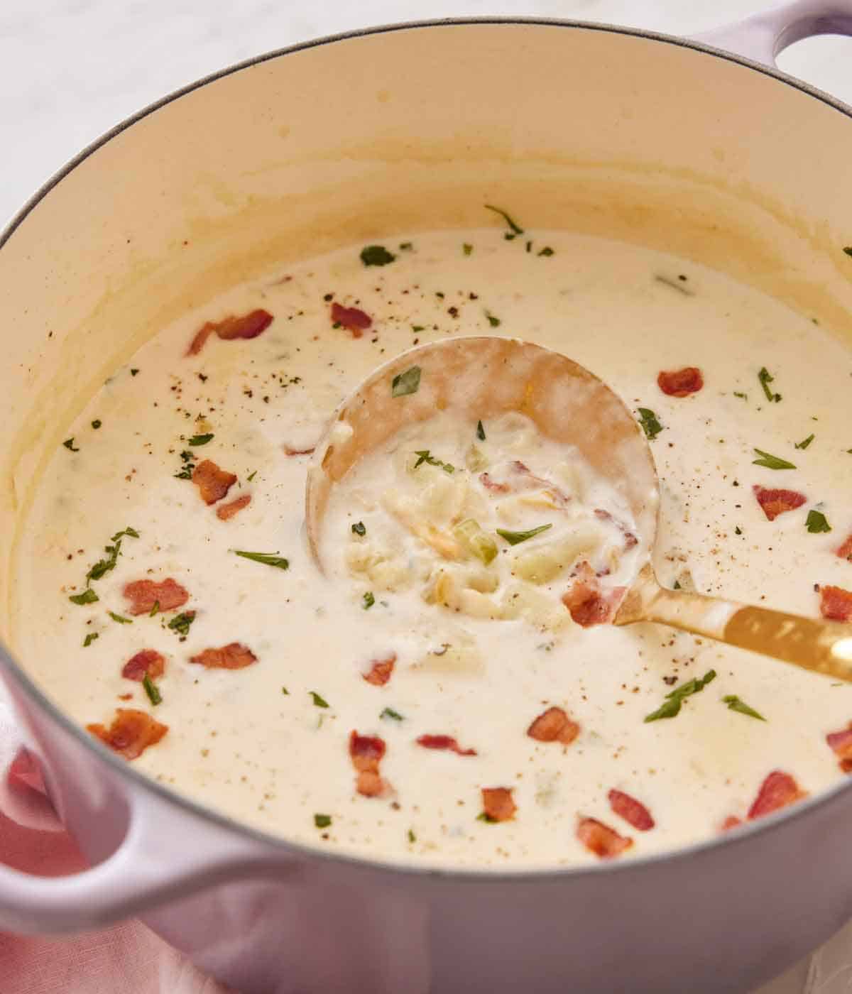 A purple dutch oven containing clam chowder with a ladle inside the soup.