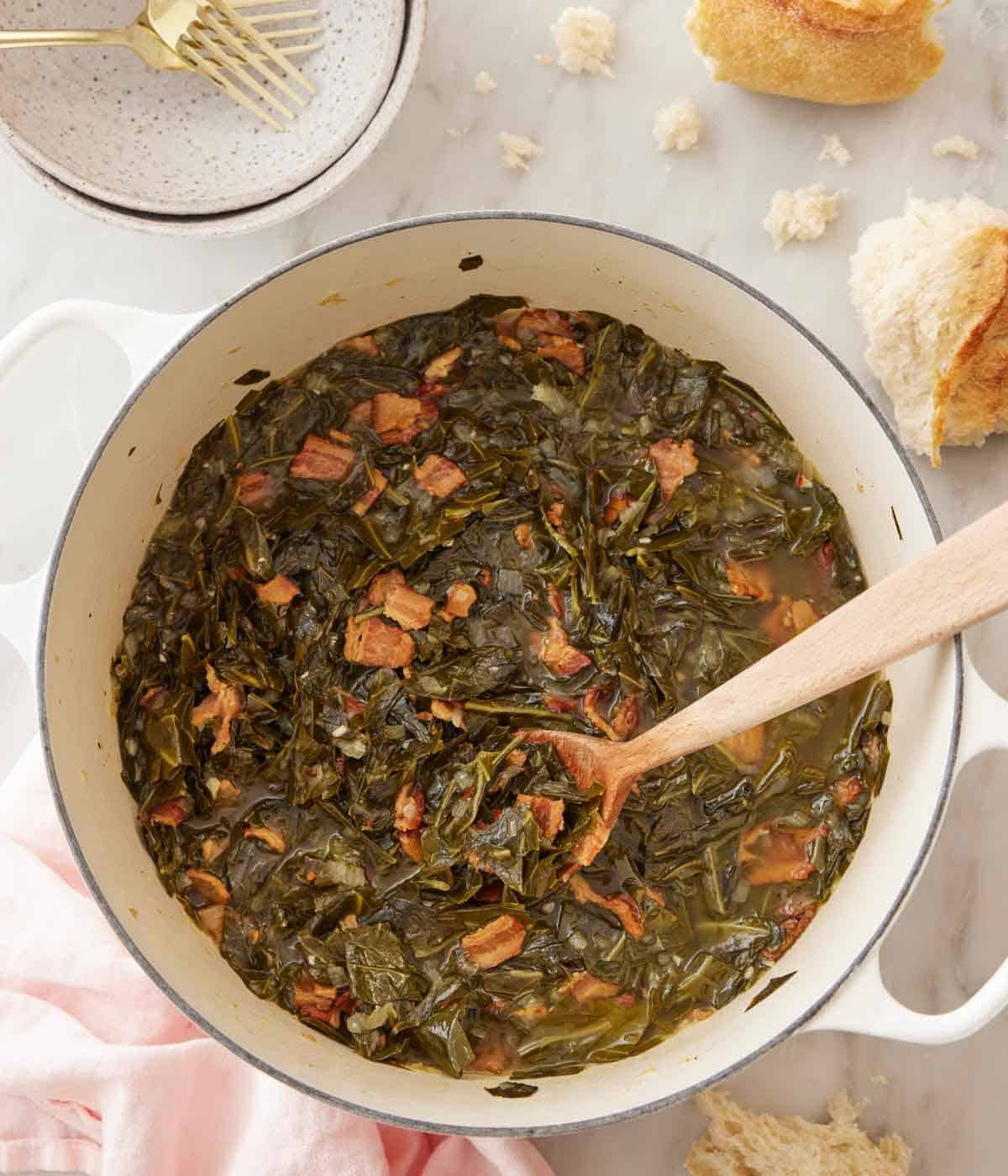 Overhead view of a pot of collard greens with a wooden spoon inside. Torn bread off to the side.