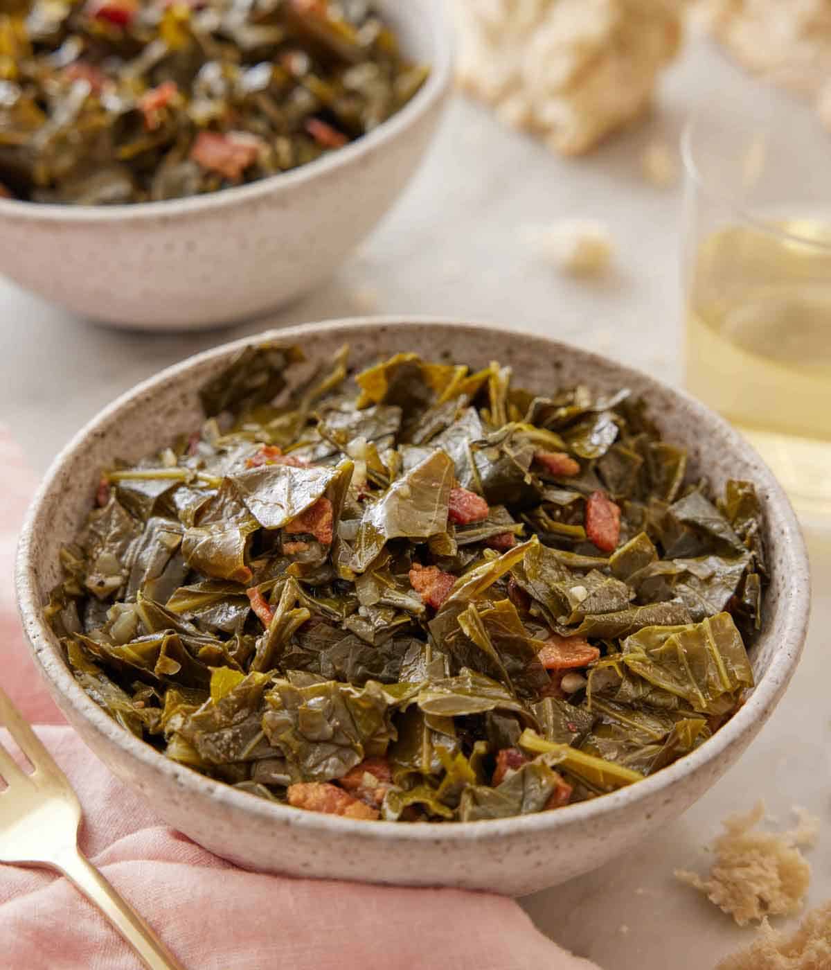 Close up view of a bowl of collard greens with a second bowl, glass of wine, and some torn bread in the back.