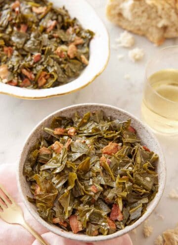 A bowl of collard greens with a serving platter and glass of wine in the background.