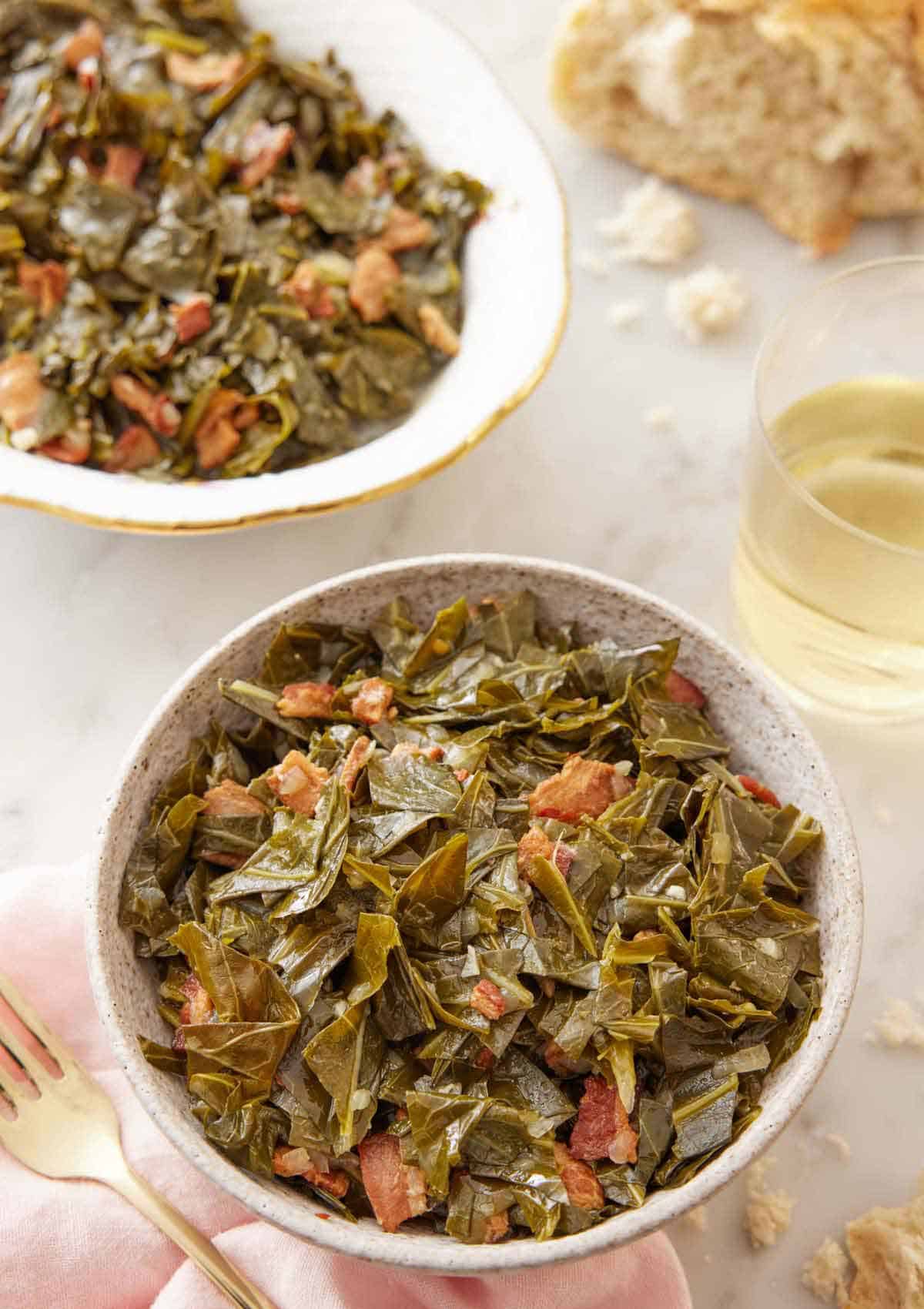 A bowl of collard greens with a serving platter and glass of wine in the background.