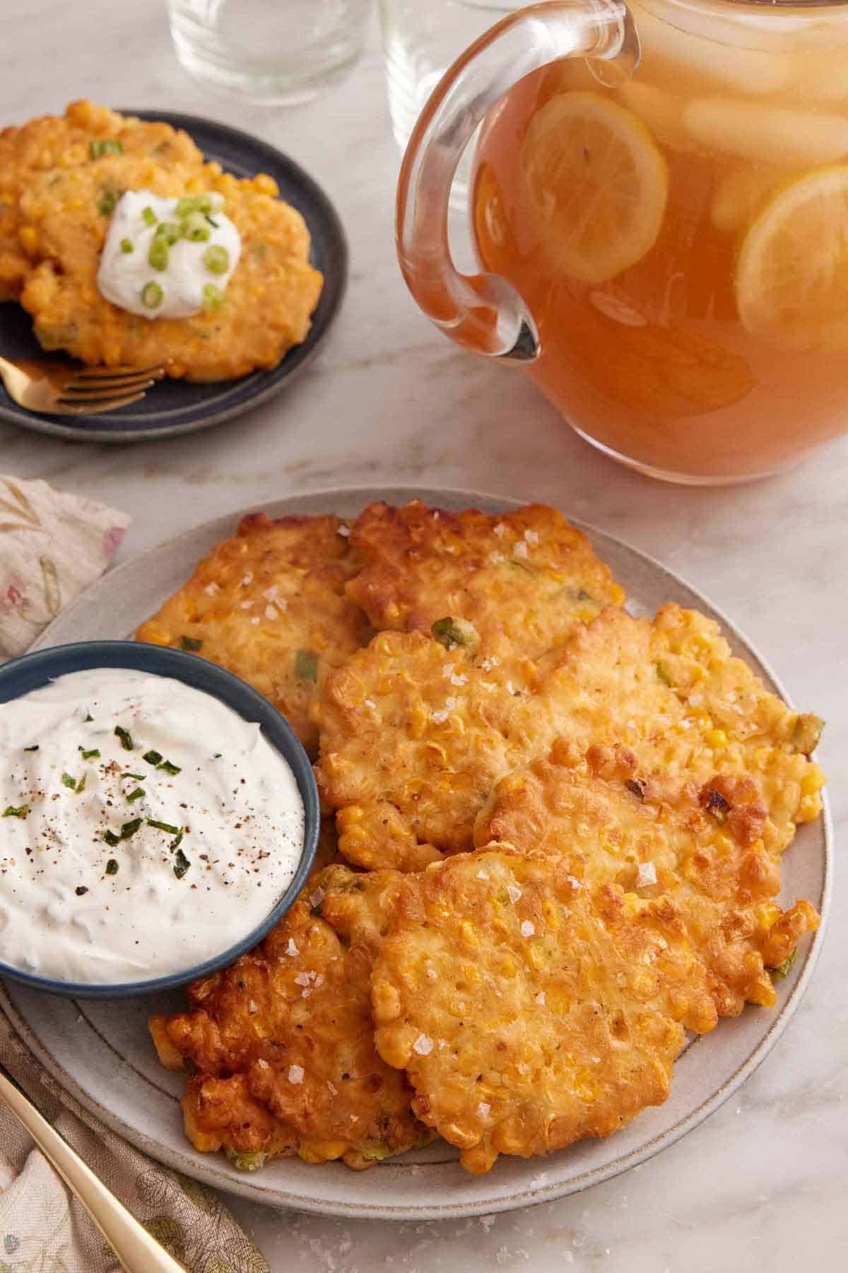 A plate with corn fritters with a bowl of dip on the side with a pitcher of iced lemon tea on the background. A serving plate with two fritters in the back.