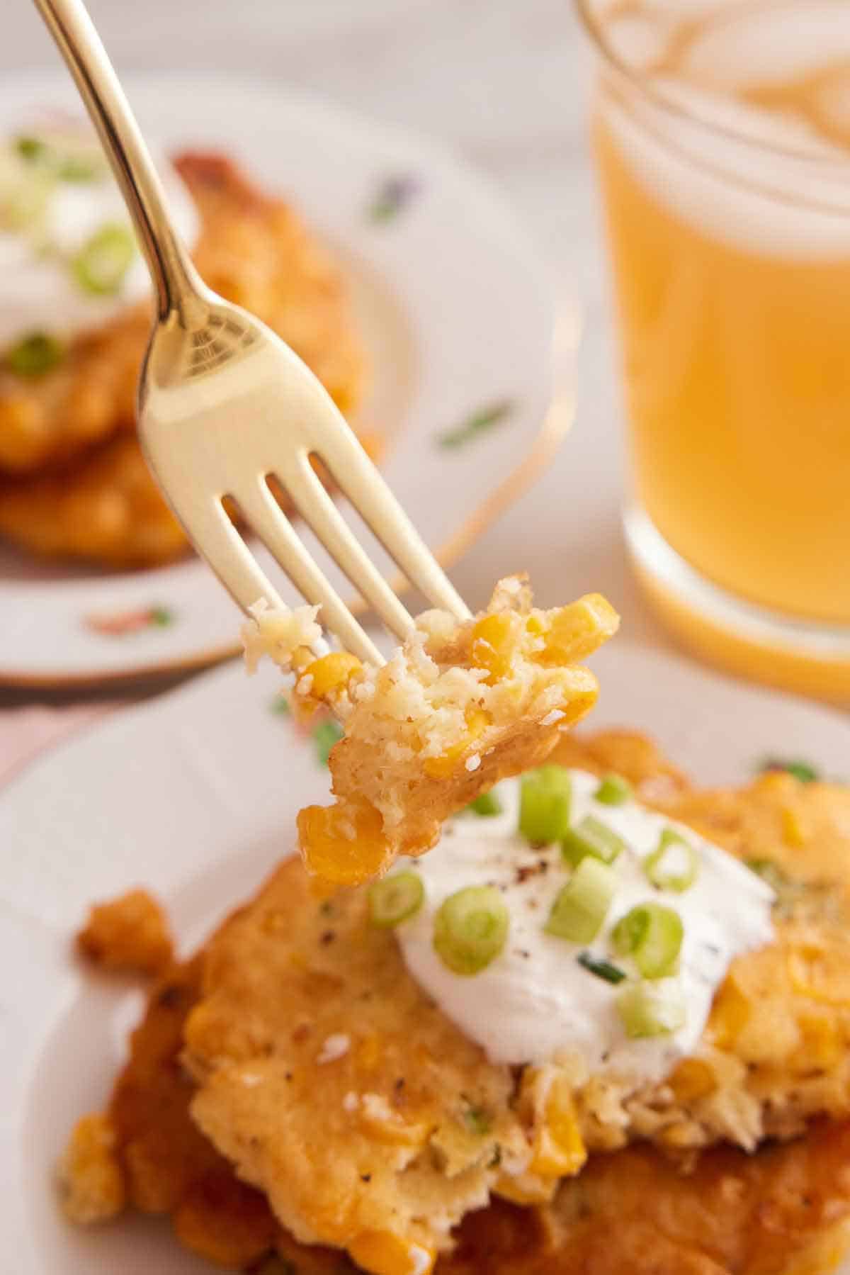 A fork lifting up a bite of corn fritters from a plate of fritters topped with sour cream and green onions.