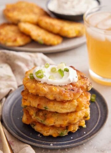 A plate with a stack of four corn fritters with sour cream on top and garnished with green onions. An iced drink in the back along with a platter of additional corn fritters.