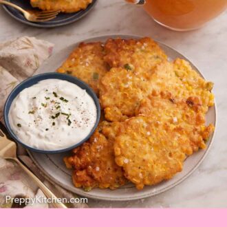 Pinterest graphic of a plate with corn fritters with a bowl of dip on the side with a pitcher of iced lemon tea on the background. A serving plate with two fritters in the back.