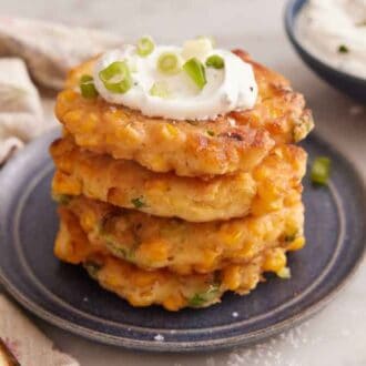 A plate with a stack of four corn fritters topped with sour cream and green onions. A bowl of dip in the back.