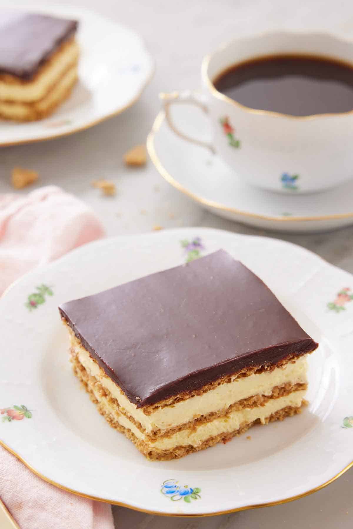 A plate with a square slice of éclair cake with a cup of coffee in the back.