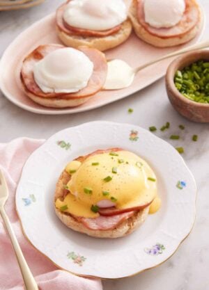 A plate with eggs benedict with hollandaise sauce over top with some chives. More eggs benedicts in the background.