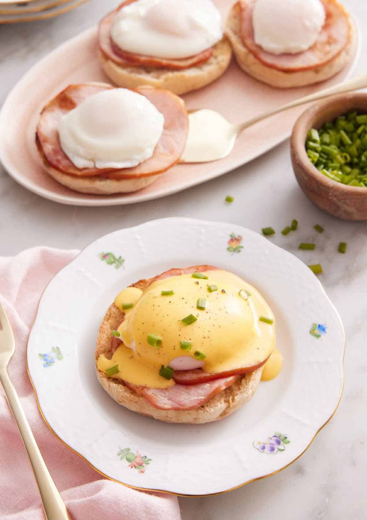 A plate with eggs benedict with hollandaise sauce over top with some chives. More eggs benedicts in the background.