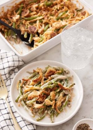 A plate with a serving of green bean casserole with a glass of water and baking dish in the background.