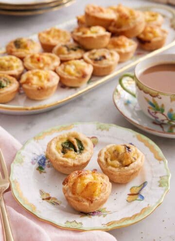 A plate with three mini quiches with a cup of coffee and a platter with additional quiches in the background.