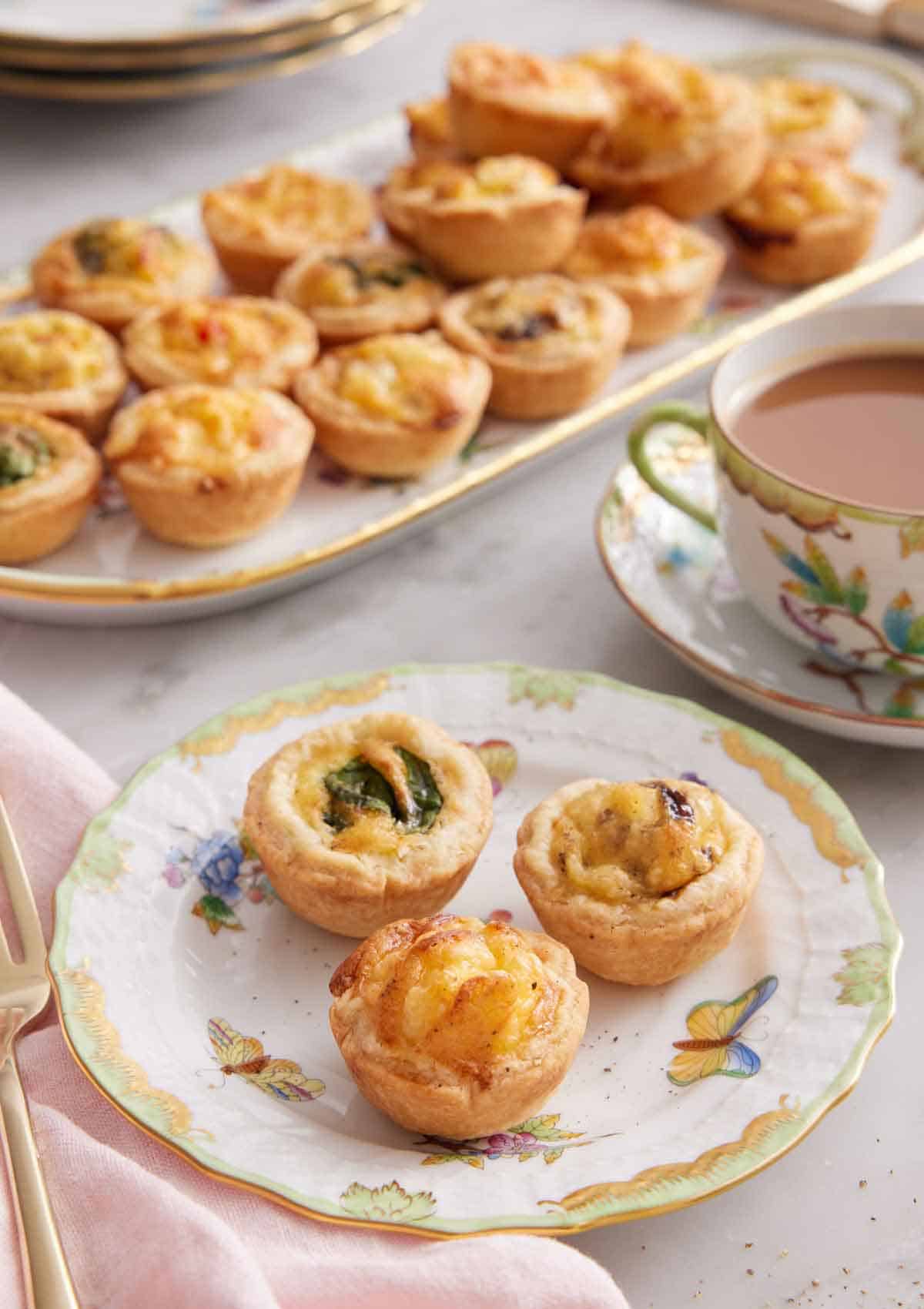 A plate with three mini quiches with a cup of coffee and a platter with additional quiches in the background.
