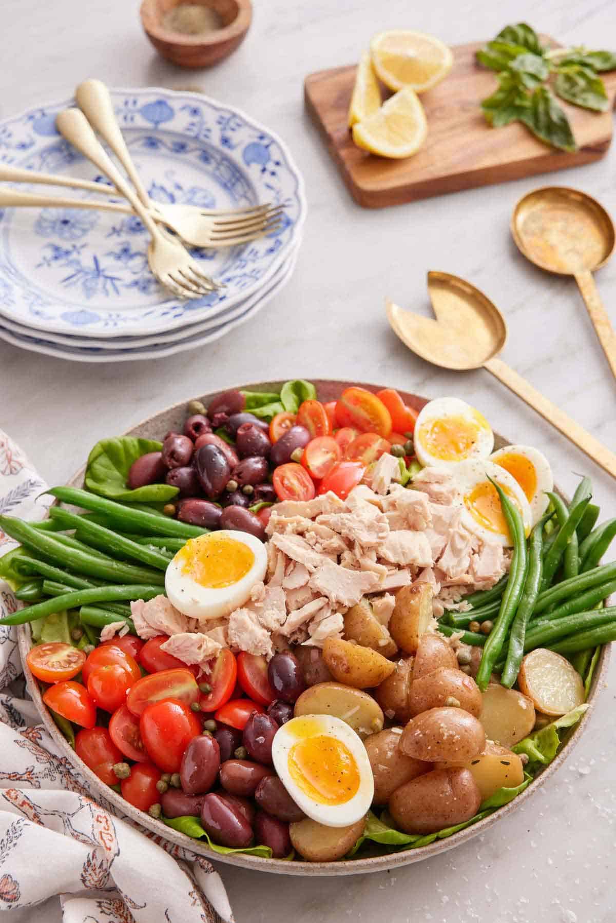 Ingredients for niçoise salad in a platter. Serving spoons, stack of plates, and forks in the background.