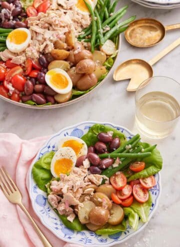 A plate of niçoise salad with a glass of wine in the back with a platter of more niçoise salad.