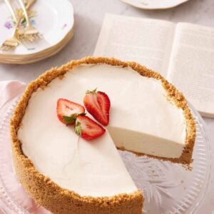 A cake stand with a no bake cheesecake with three pieces of strawberries on top with a slice cut out. An opened book and slice of cake in the background with some plates and forks.