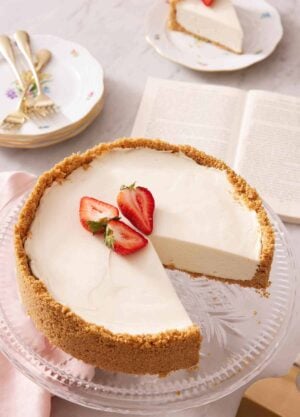 A cake stand with a no bake cheesecake with three pieces of strawberries on top with a slice cut out. An opened book and slice of cake in the background with some plates and forks.