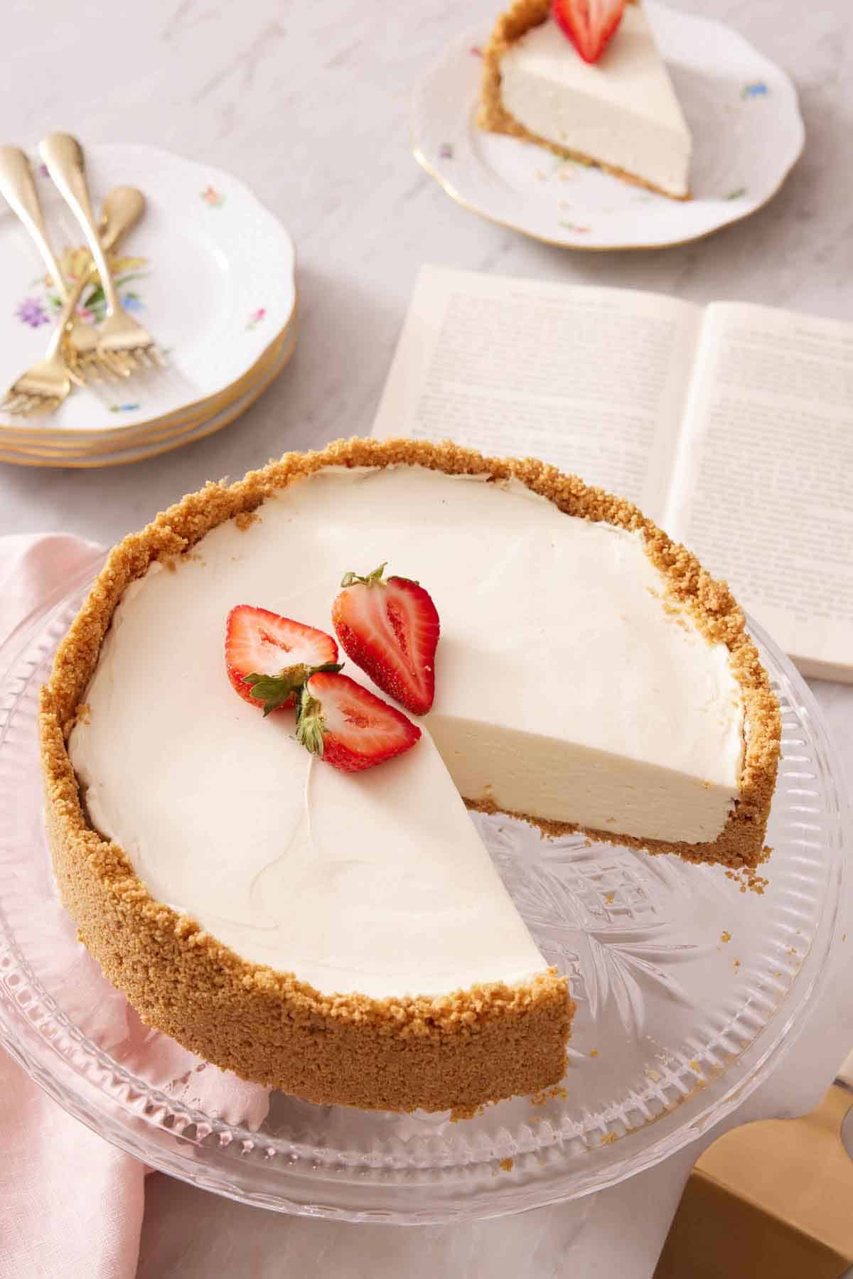 A cake stand with a no bake cheesecake with three pieces of strawberries on top with a slice cut out. An opened book and slice of cake in the background with some plates and forks.