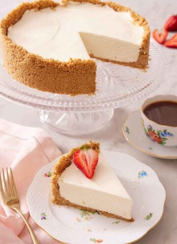 A plate with a slice of no bake cheesecake with a piece of strawberry on top with a cake stand with the rest of the cake.