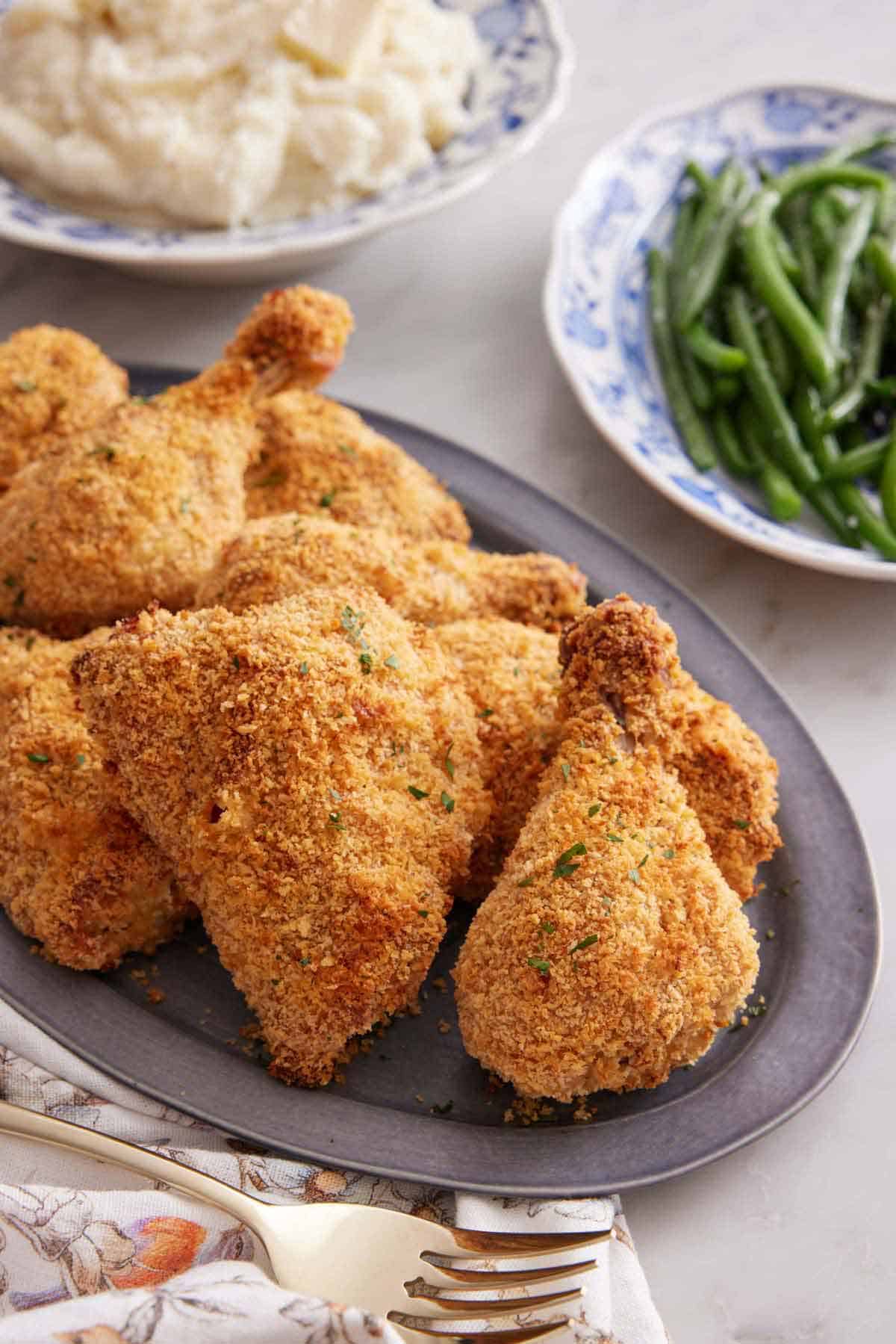A platter of oven fried chicken with plates of green beans and mashed potatoes in the background.