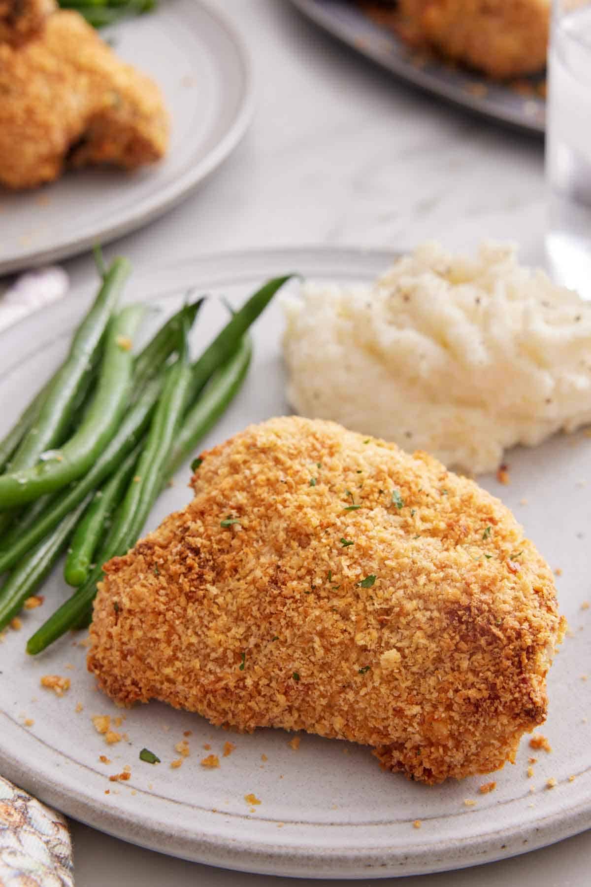 A oven fried chicken thigh on a plate with some green beans and mashed potatoes.