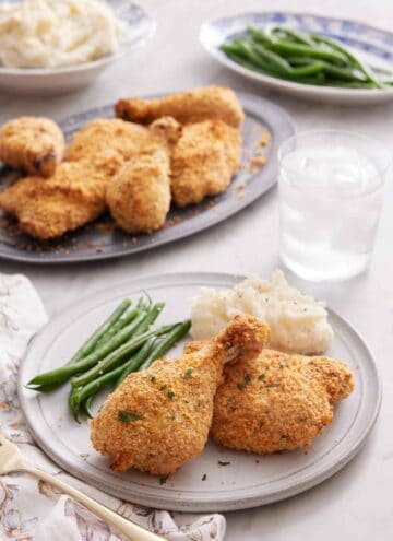A plate of oven fried chicken with green beans and mashed potatoes. A glass of water and platters of food in the background.
