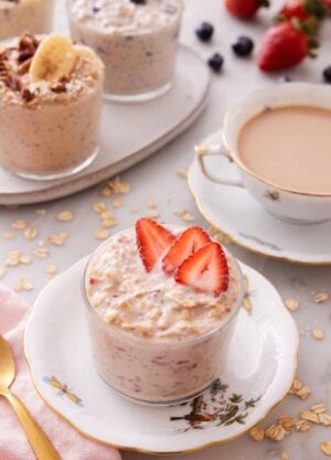 A plate with a glass of overnight oats topped with strawberries with a cup of coffee and more overnight oats in the background.