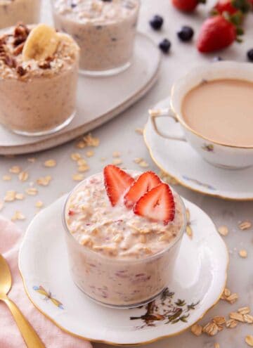 A plate with a glass of overnight oats topped with strawberries with a cup of coffee and more overnight oats in the background.