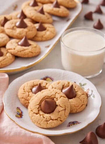 A plate with three peanut butter blossoms with a glass of milk and a platter of more peanut butter blossoms in the background.