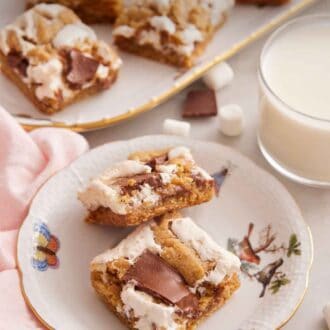 Pinterest graphic of a plate with two pieces of s'mores bars with a cup of milk and platter of bars in the background.