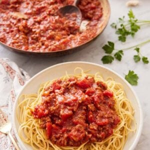 A bowl of noodles with spaghetti sauce on top with a skillet full of sauce in the background.