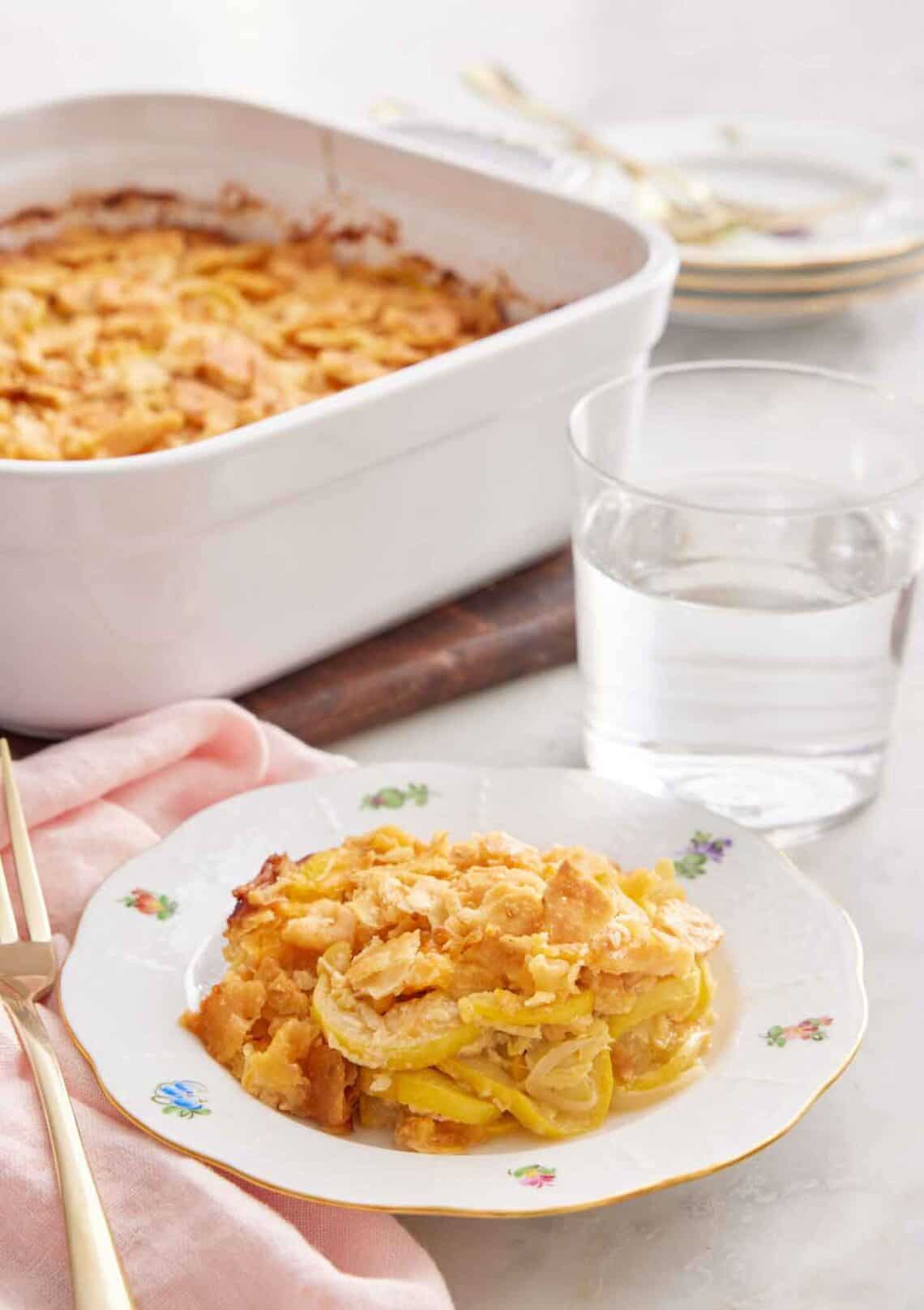 A plate with a serving of squash casserole with a glass of water and baking dish in the background.