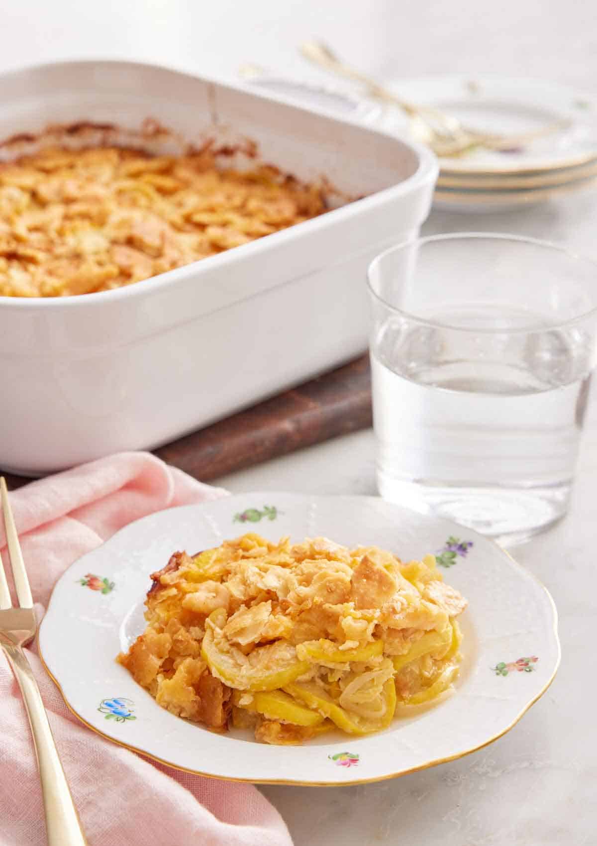 A plate with a serving of squash casserole with a glass of water and baking dish in the background.