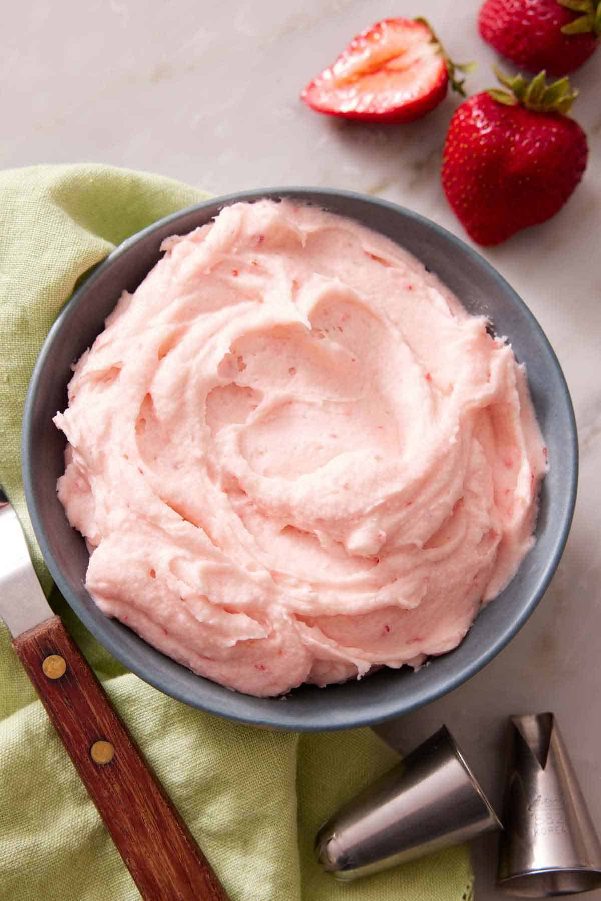 A bowl of strawberry frosting with fresh strawberries on the side and piping tips.