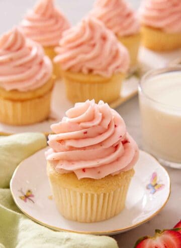 A vanilla cupcake with strawberry frosting piped on top on a small plate with additional frosted cupcakes in the background.