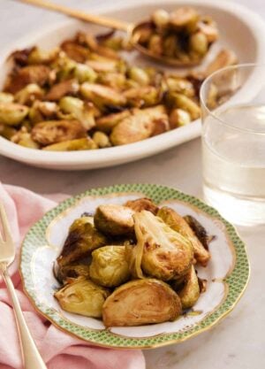 A bowl of air fryer Brussels sprouts with a drink and platter of more sprouts in the background.