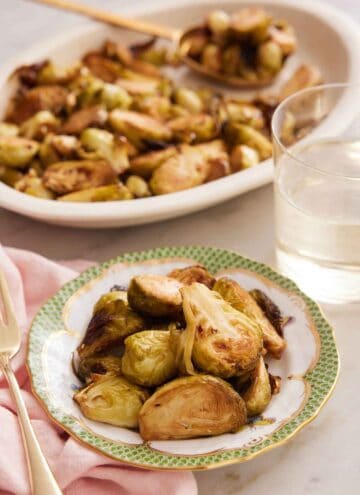 A bowl of air fryer Brussels sprouts with a drink and platter of more sprouts in the background.