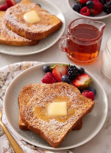 A plate of air fryer french toast with powdered sugar, butter and syrup on top with berries on the side. A small vessel of syrup and another plated serving in the background.